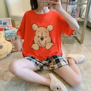 Zweiteiliger Pyjama mit T-Shirt, Winnie-Muster und modischen Karo-Shorts, getragen von einer Frau, die auf einem Karo in einem Haus sitzt