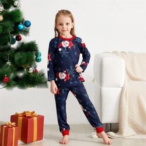 Pyjama mit Schneeflockendruck und Weihnachtsmann für die Familie, getragen von einem kleinen Mädchen vor einem Weihnachtsbaum in einem Haus