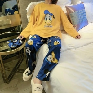 Netter Disney Donald Pyjama für Frauen in Senfgelb und Blau, der von einer Frau getragen wird, die auf einem Bett in einem Haus sitzt