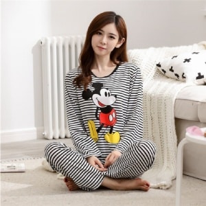 Gestreifter Mickey-Mouse-Pyjama für Frauen, der von einer Frau getragen wird, die in einem Haus vor einem Bett sitzt