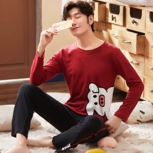 Baumwollpyjama mit rotem Pullover mit Schweinchenmuster und schwarzer Hose, getragen von einem Mann, der auf einem Teppich in einem Haus sitzt