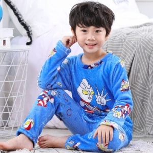 Blauer Flanellpyjama mit Superhelden-Motiv für blaue Jungen, getragen von einem kleinen Jungen, der auf einem Teppich vor einem Bett in einem Haus sitzt