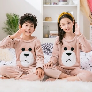 Brauner Kinderpyjama aus Flanellfleece, getragen von einem kleinen Mädchen und einem kleinen Jungen, die auf einem Teppich in einem Haus sitzen