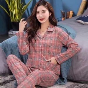 Modischer zweiteiliger karierter Pyjama mit Umlegekragen für Frauen, der von einer Frau getragen wird, die auf einem Teppich vor einem Bett in einem Haus sitzt