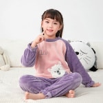 Polarpyjama-Set für Kinder in lila, getragen von einem kleinen Mädchen, das auf einem Bett in einem Haus sitzt