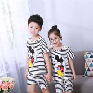 Schwarz-weiß gestreifter Sommerpyjama mit Mickey-Mousse-Aufdruck für Kinder, der von einem kleinen Jungen und einem kleinen Mädchen vor einem Stuhl in einem Haus getragen wird
