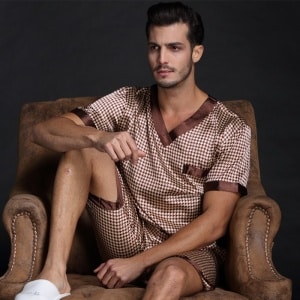 Kurzärmeliger, karierter Pyjama aus Seidensatin für Männer in braun mit einem Mann, der den Pyjama auf dem Sofa trägt