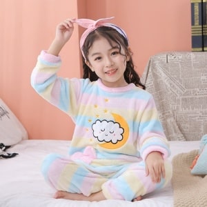 Regenbogenfarbener Fleece-Pyjama für Kinder, der von einem kleinen Mädchen auf einem Bett in einem Haus getragen wird