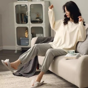 Sommerpyjama mit weißem Pullover, Fledermausärmeln und grauer Hose, getragen von einer Frau, die auf dem Sofa in einem Haus sitzt