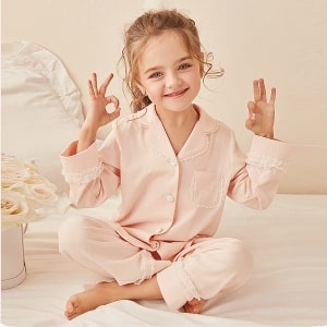 Zweiteiliger Pyjama aus Spitze für "doing as Mommy" mit einem kleinen Mädchen, das den Pyjama trägt, mit einem weißen Kinderbett und einem rosa Hintergrund
