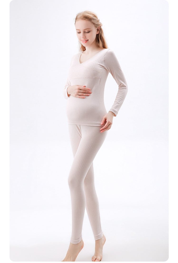schwangere Frau, die sich mit einer Hand am Bauch berührt, trägt einen beigen Pyjama, der aus einer Hose und einem langärmligen Pullover besteht
