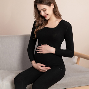 eine junge schwangere Frau sitzt in einem schwarzen Schwangerschaftspyjama und lächelt, während sie ihren runden Bauch berührt